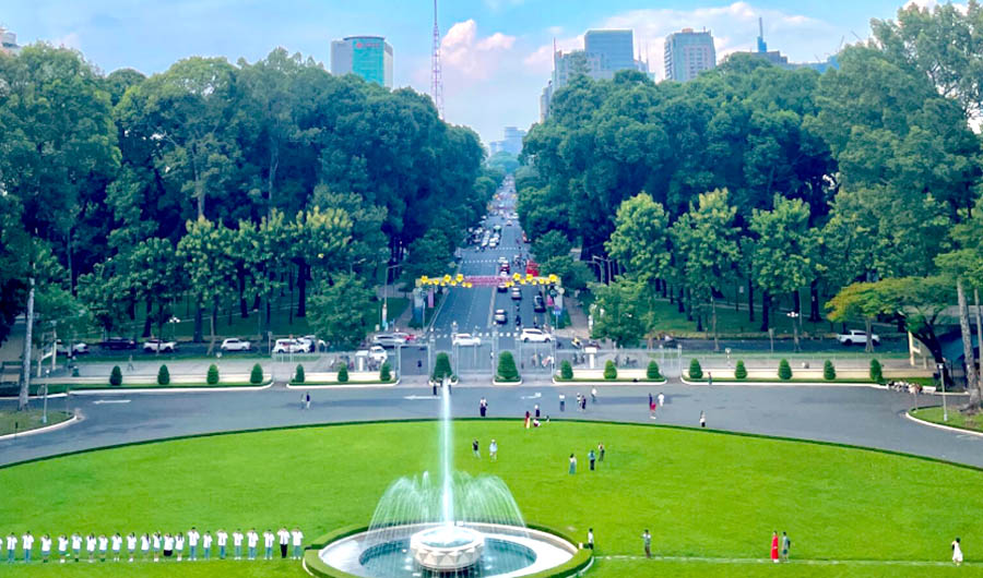 A fountain in a park with trees and buildings in the backgroundDescription automatically generated